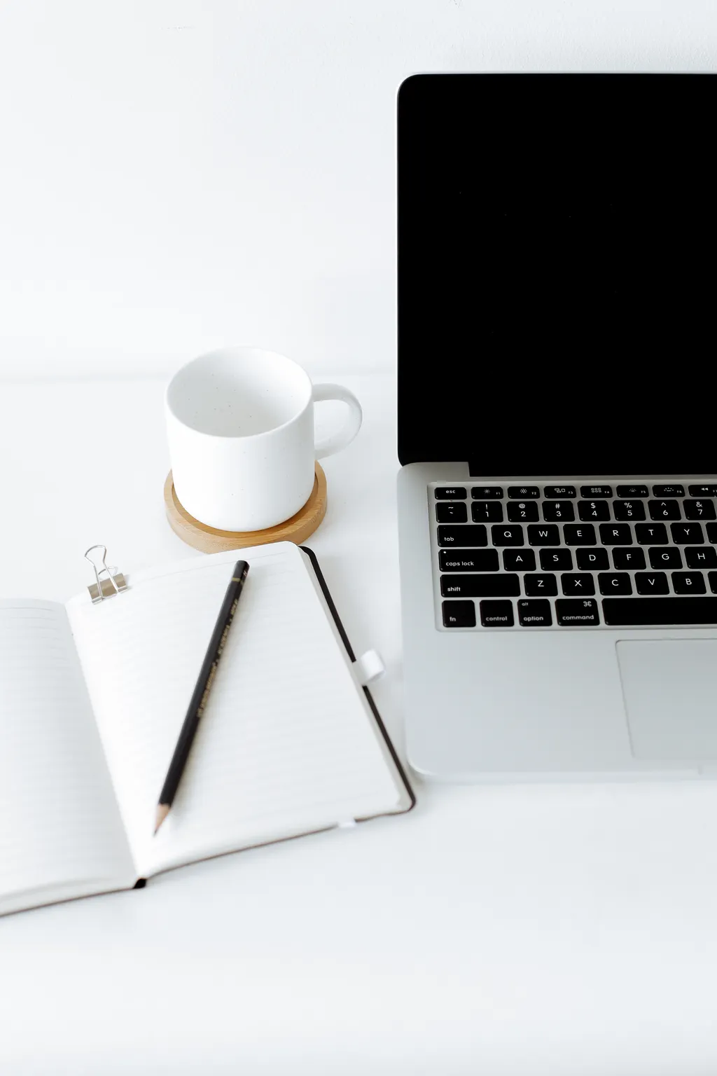 An open notebook and pencil beside a laptop and a coffee cup on a white desk